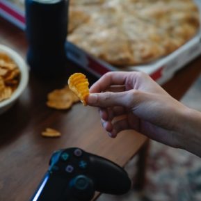 hand holding crisp in one hand and games console in another with pizza box in the background