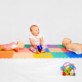 Baby sitting on a coloured mat next to two first aid dolls
