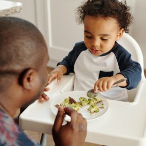 Man sitting in front of a young child while he eats from his highchair